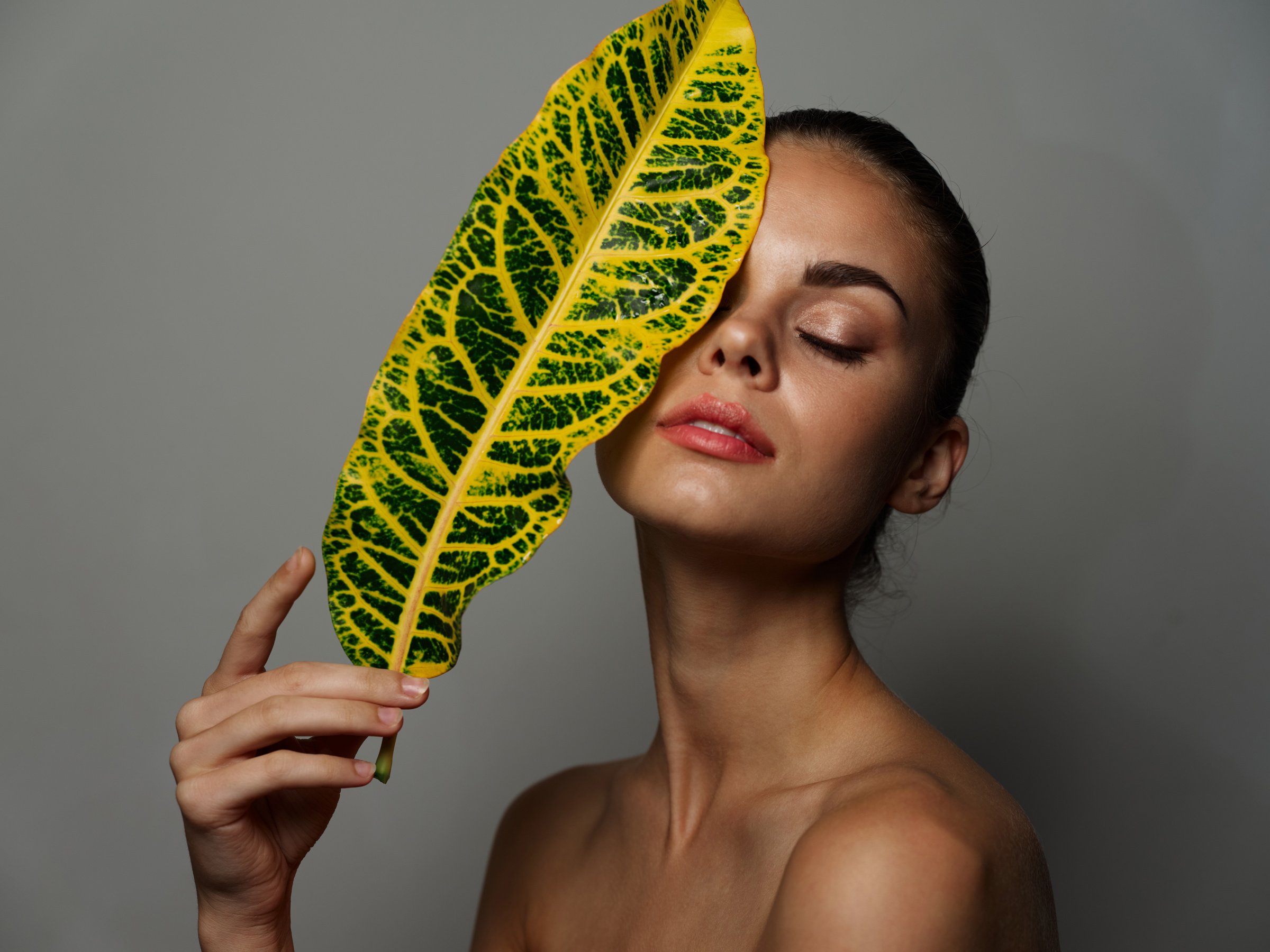 Beautiful Woman with Closed Eyes and Green Leaf in Front of Face on Gray Background